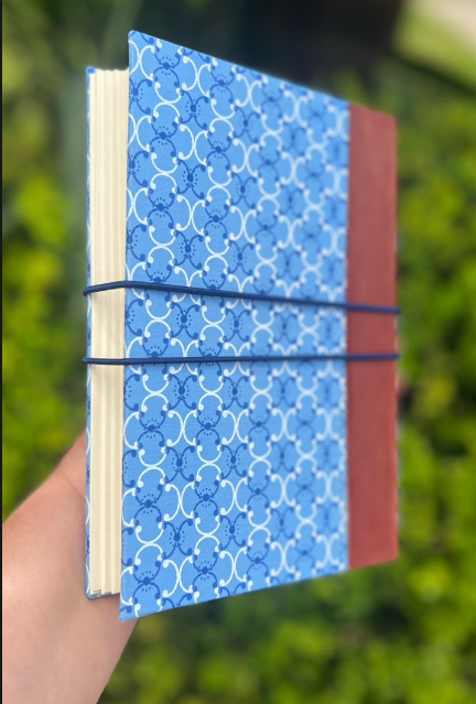 Caderno artesanal azul visto em perspectiva, com lombada vinho e elásticos de fechamento